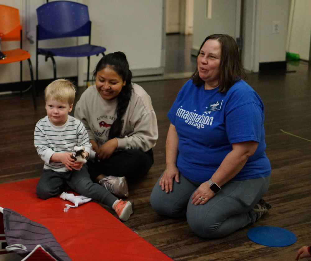 3 people sit on the floor. Far left a blonde haired toddler sits very close to a dark haired woman, she is leaning close to the child. A woman in a blue Imagination Stage shirt sits on her knees, to their right. 